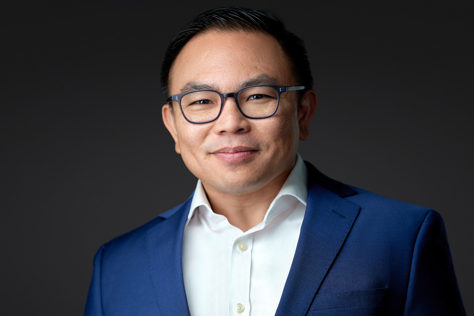 Professional headshot of a man wearing glasses and a navy blazer, dark studio background