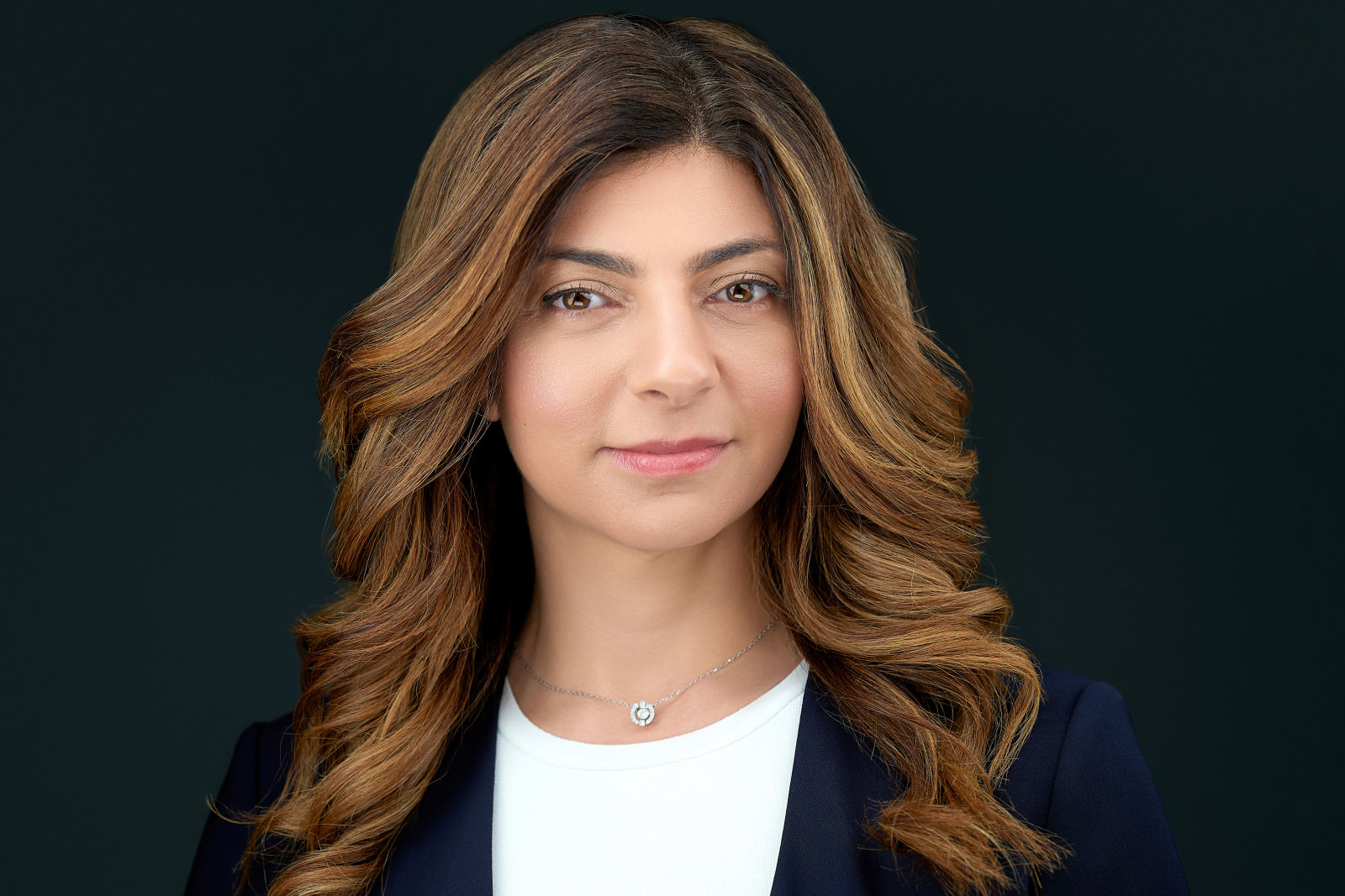 Professional headshot of a woman with long wavy brown hair wearing a navy blazer, dark studio background