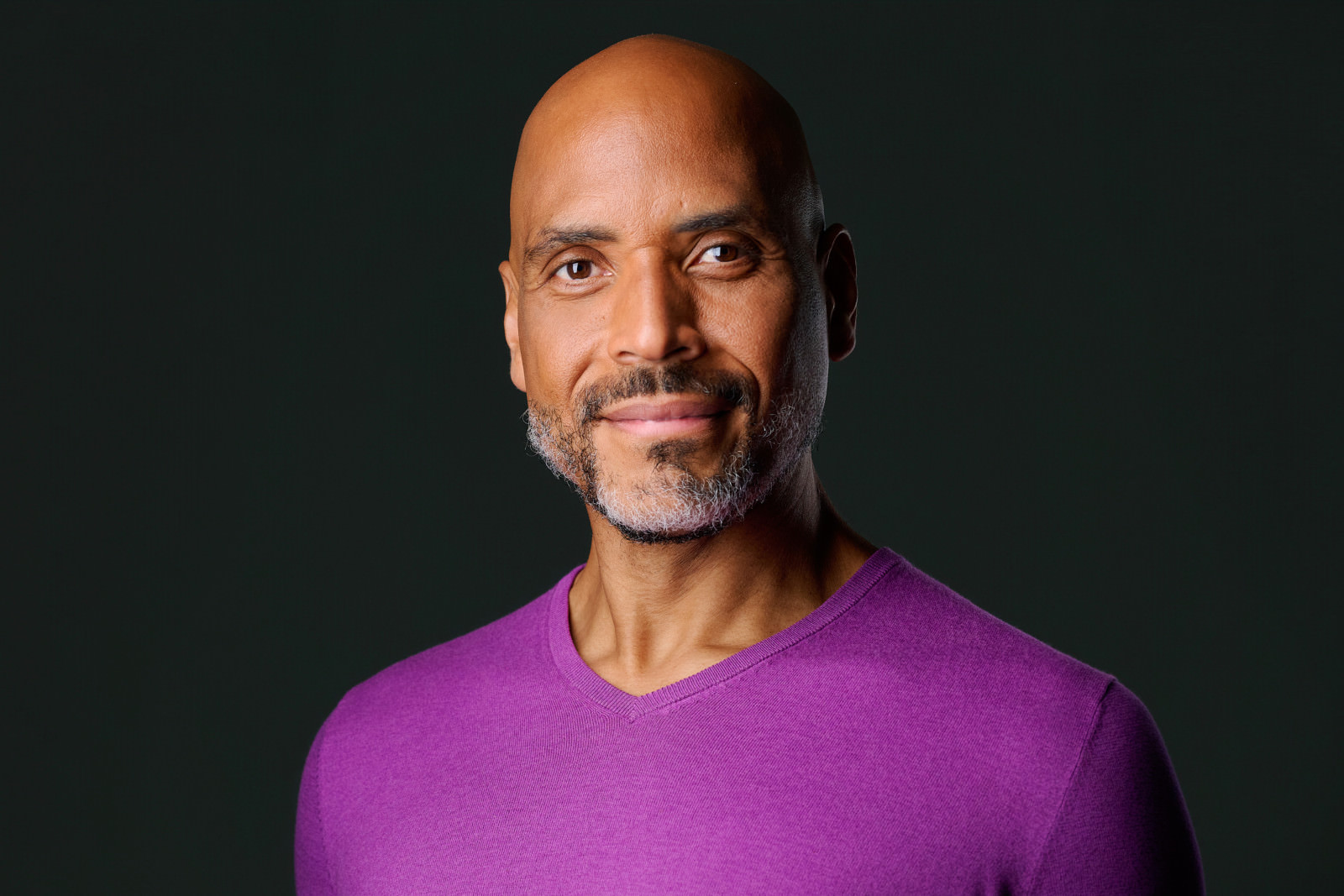 Professional headshot of a bald man with a gray beard wearing a purple sweater, dark studio background